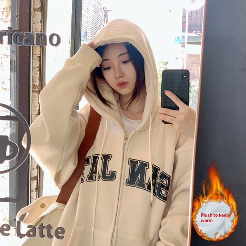 Woman wearing a cream zip-up loose casual-style hoodie with varsity lettering, taking a mirror selfie.