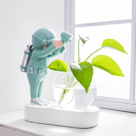 Astronaut gold statue and green diver resin figures with a round glass hydroponic flowerpot on a white base.