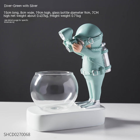 Green diver figurine with silver gear leaning over a round glass hydroponic flowerpot on a white base.