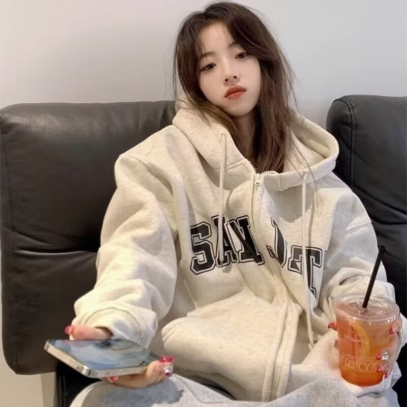 Woman in zip-up loose casual-style hoodies, oversized light gray with varsity letters, on couch with phone.