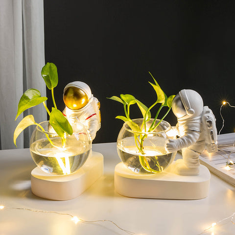 Two resin glass hydroponic flowerpots: a gold astronaut and a green-diver figurine on white bases with glowing plants.