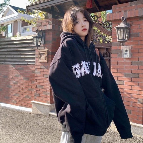 Young woman in an oversized black zip-up hoodie with white SAJA lettering beside a brick wall.